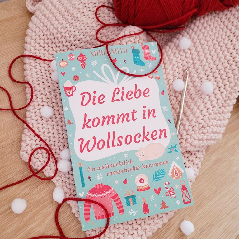 Das Buch Die Liebe kommt in Wollsocken liegt auf einer kuscheligen rosa Strickdecke, daneben Strickzeug.