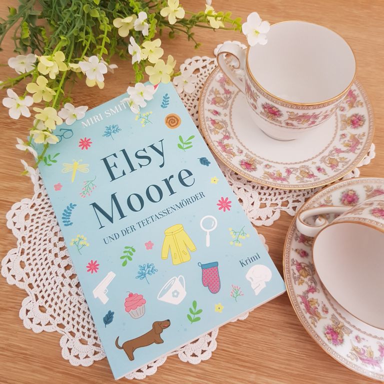 Das Buch Elsy Moore und der Teetassenmörder, daneben Blumen und Teetassen.